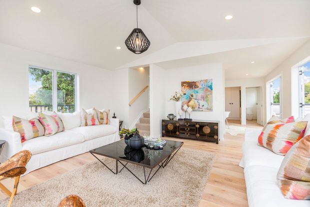Bright, airy living room with white sofas, colourful patterned cushions, a black glass coffee table, textured area rug, pale timber flooring, a built-in dark timber fireplace cabinet, and a sculptural pendant light. Large windows provide views to greenery, while a stairway and hallway are visible in the background.