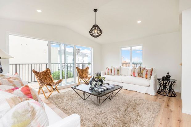 Bright contemporary living room with white walls and sofa seating, patterned cushions, woven lounge chairs, a black geometric coffee table, pendant light, light timber flooring, and large glazed doors opening to a balcony. The room has abundant natural light and a relaxed, airy feel.