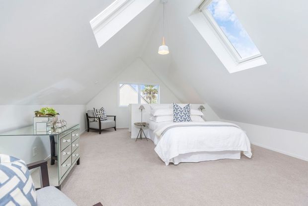 Bright attic-style bedroom with a steep sloped ceiling, two skylights, a central pendant light, soft grey carpet, white walls, and a white upholstered bed. The room includes mirrored drawer storage, a small armchair, bedside tables, and a compact rear window with greenery visible outside.