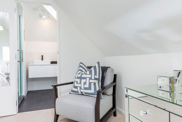 Bright, compact attic-style sitting area with a sloped white ceiling, a mid-century-inspired upholstered armchair, and a mirrored glass-front chest. An open doorway reveals a contemporary bathroom with a white vanity, glass shower enclosure, and dark floor tiles.
