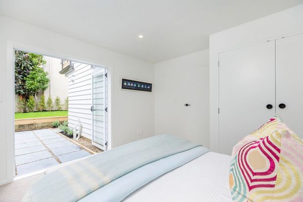 Bright, compact bedroom with white walls and built-in white storage cupboards. The bed is dressed in pale blue and white bedding with a colourful patterned accent pillow. An open glazed door provides views to a landscaped courtyard and paved outdoor path, while a small framed artwork adds a subtle decorative element.