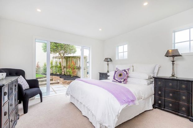 Bright, generously sized bedroom with a white upholstered bed dressed in white linens and a lavender-purple throw and accent cushion. Dark traditional timber dressers and bedside tables, classic lamps, a deep charcoal armchair, recessed ceiling lights, pale carpet, and white walls create a refined, softly traditional interior. Large glazed doors open to a small landscaped courtyard.