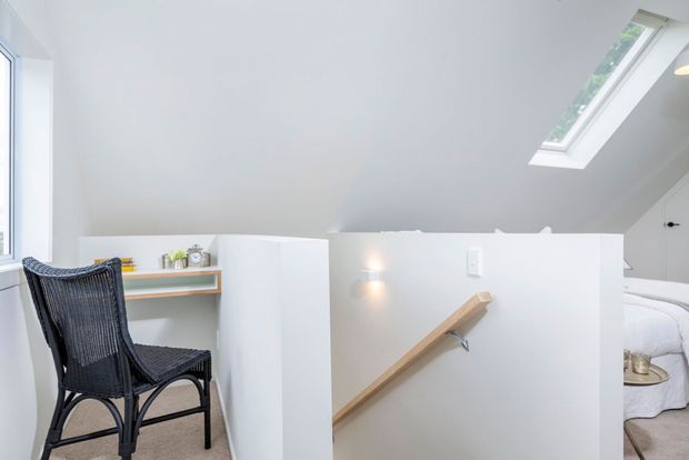 Bright, compact attic landing or stairway with white walls and a sloped ceiling, featuring a skylight, a timber handrail, and a small built-in desk with a black woven chair. A partially visible bed through the opening suggests an adjoining bedroom.