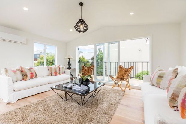 Bright contemporary living room with white slipcovered sofas, colourful patterned cushions, a glass coffee table with a geometric black metal base, a textured neutral rug, and two woven sling chairs. Large windows and glazed doors open toward a balcony, creating an airy indoor-outdoor connection. A black geometric pendant light and recessed ceiling lights provide simple decorative detailing.