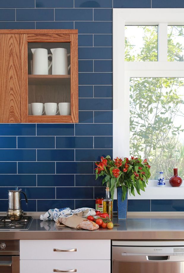 A compact, richly coloured kitchen vignette with glossy dark-blue subway tile walls, warm timber wall cabinetry with an open display section, white lower drawers, and a stainless-steel benchtop and dishwasher. A large white-trimmed window brings in natural light, while countertop items and a leafy flowering plant add lived-in character.