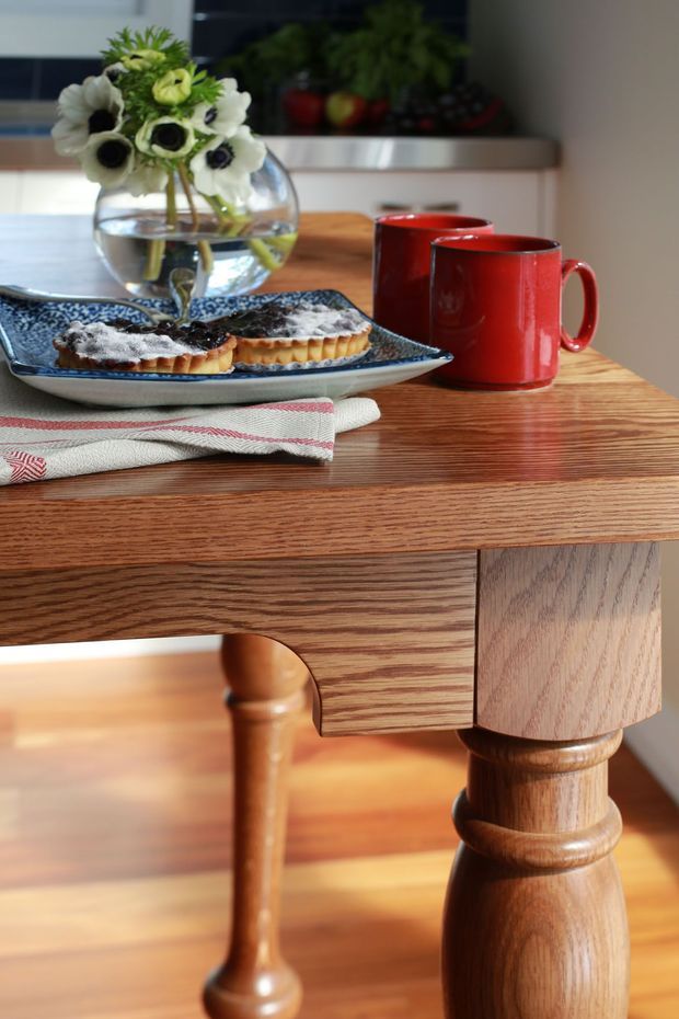 Close-up interior still life of a traditional timber dining table with turned legs and a carved apron. The table is styled with two red mugs, a blue patterned plate holding pastries, a folded white-and-red tea towel, and a glass vase of flowers. Warm timber flooring and a light neutral kitchen or dining background are visible.