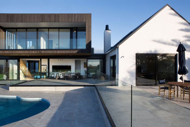Exterior view of a contemporary residential courtyard centered on a swimming pool and paved terrace. The home combines a dark timber-clad, glazed two-storey wing with a white rendered gable-roofed volume. Floor-to-ceiling glazing reveals an open-plan living area and internal stair, while outdoor dining furniture sits beside the pool.