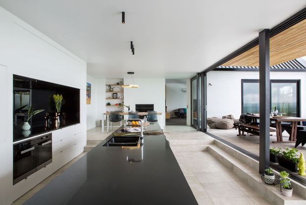 Open-plan contemporary kitchen and living area with a long dark island, integrated black appliances, white walls, and pale stone flooring. Full-height sliding glazing opens to a covered outdoor dining and lounge area, with timber ceiling lining and dark structural framing. The room has a bright, refined indoor-outdoor layout with minimal fixed detailing.