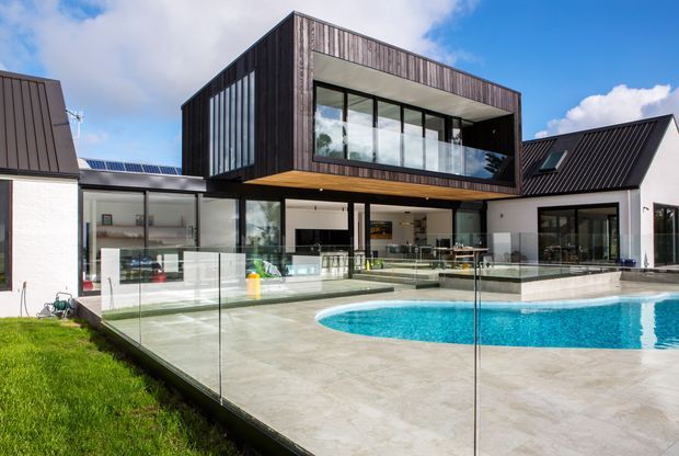 A contemporary multi-level residence with a dramatic cantilevered upper volume clad in dark vertical timber, large glazed openings, and adjoining pitched-roof wings. The ground floor opens onto a broad paved terrace and bright blue swimming pool enclosed by frameless glass balustrading. The composition features strong geometric forms, dark metal roofing, extensive glazing, and a clean indoor-outdoor connection under a partly cloudy sky.
