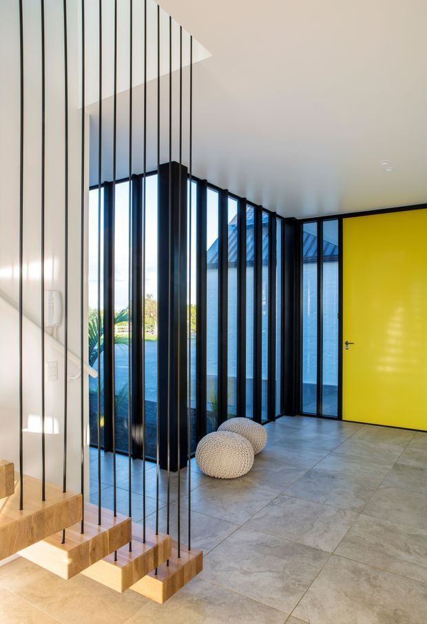 A contemporary residential entryway with a floating timber staircase suspended on slender black metal rods. Full-height black-framed glazing lines the corridor, bringing in abundant daylight, while a vivid yellow door creates a strong focal point. Large-format grey floor tiles, white walls and ceiling, and knitted round stools complete the clean, graphic interior.