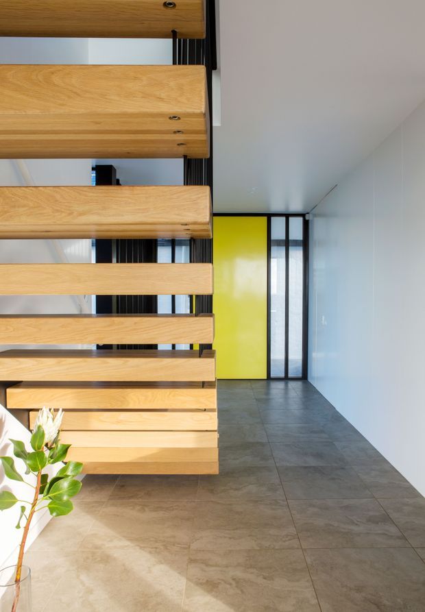 Bright contemporary interior hallway featuring a sculptural open-riser timber staircase with thick horizontal oak treads and black vertical supports. The staircase partially screens a corridor leading to a vivid lime-yellow door and black-framed glazed opening. Large grey stone-look floor tiles, white walls and ceiling, and a small floral arrangement create a crisp modern composition.