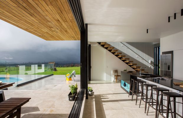 A contemporary open-plan kitchen and dining area opening onto a large covered terrace and poolside entertaining space. Floor-to-ceiling glazed sliding doors connect the interior and exterior, while a sculptural open staircase with dark vertical balustrading leads upward. The kitchen features a long dark island with bar seating, pale stone-look flooring, white walls and ceiling, and strong natural light. A timber-lined soffit extends over the terrace, with expansive rural green views and dramatic clouds beyond.