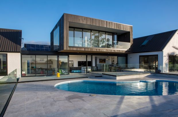 Exterior view of a large contemporary residence arranged around a swimming pool. The home combines a dark pitched-roof wing with a projecting upper-level timber-clad volume, extensive floor-to-ceiling glazing, and a broad paved pool terrace. Interior living areas are visible through the glazing, with a glass balustrade along the terrace edge.
