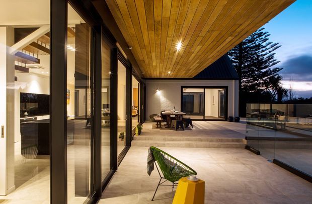A contemporary covered outdoor terrace opening directly from a glazed interior living space. The terrace features a large timber-lined soffit with recessed spotlights, pale stone or concrete-look paving, frameless glass balustrades, and an outdoor dining setting. A dark pitched roof edge and extensive black-framed glazing define the architecture, with a pool or lower outdoor area visible beyond the balustrade. The interior kitchen or living area is visible through the open glazing.