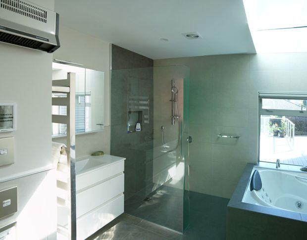 Modern bathroom with a glass-enclosed walk-in shower, wall-mounted shower fittings, white floating vanity, large grey wall and floor tiles, and a built-in spa bath beside a window. A bedroom or adjoining space is partially visible through the open doorway at left.