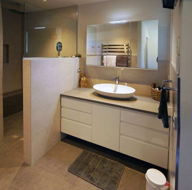 Contemporary bathroom featuring a frameless glass shower enclosure, tiled walls and floor, a wall-mounted light-toned vanity with integrated drawers, a pale stone-look benchtop, and a white vessel basin with chrome tapware. A large mirrored cabinet spans the vanity wall, with a heated towel rail visible in reflection. The room has warm neutral finishes and practical built-in storage.
