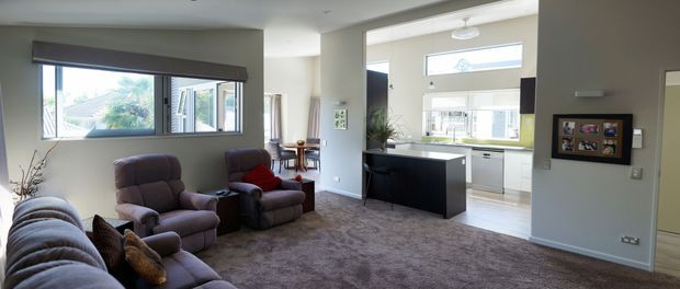 A bright open-plan living room with plush charcoal and mauve seating, a large textured rug, and a compact dining area. The room opens directly into a contemporary white kitchen with a dark island, framed by large windows and glazed doors that bring in abundant daylight.