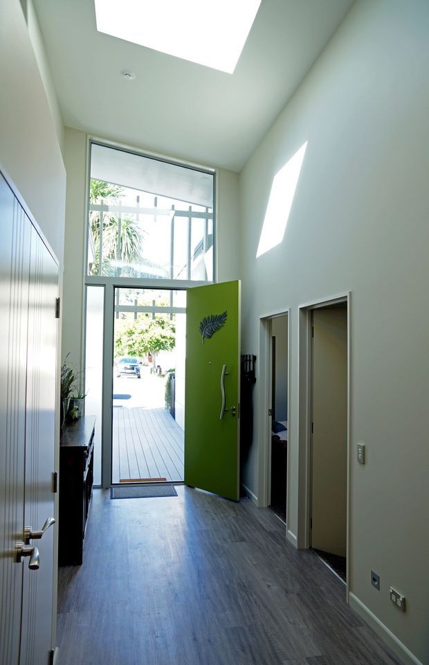 Bright contemporary residential entry hall viewed from inside, with a tall glazed front door and transom, vivid green pivot door, dark timber-look flooring, pale walls, a skylight, and side doorways. The entry opens onto a timber deck outside.