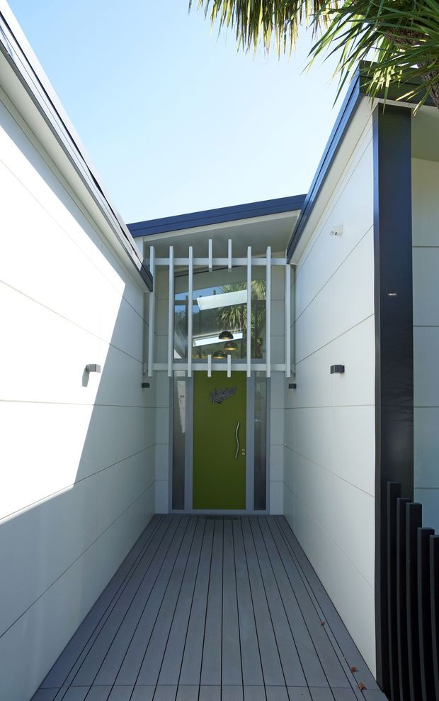 A narrow covered entry passage leading to a contemporary front door. The space has white horizontal wall cladding, a dark grey decked floor, a tall glazed entrance with vertical white fins, and a vivid green door. Strong geometric lines, dark trim, and tropical planting visible overhead create a bright modern exterior approach.