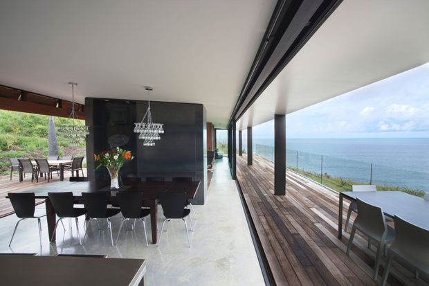 Contemporary open-plan dining area with a long dark dining table, modern black chairs, and a dramatic crystal pendant light. Full-height sliding glazing opens the room to a wide timber deck overlooking the ocean, with a second outdoor dining setting visible beyond. The space has a flat white ceiling, dark structural columns and wall cabinetry, polished pale flooring, and expansive coastal views.