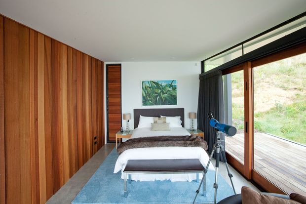 A warm, contemporary bedroom with a large bed centered against a white wall, full-height vertical timber wall panelling and built-in wardrobe cabinetry along the left side. Floor-to-ceiling timber-framed glazing opens toward a timber deck and landscaped exterior, with a high clerestory window above. The room includes bedside tables and lamps, a bench at the foot of the bed, a blue area rug, dark curtains, artwork, and a telescope. The space is bright, refined, and connected to the outdoors.
