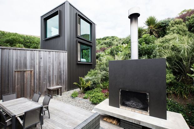 Exterior view of a compact contemporary dark-clad structure beside a timber deck and substantial outdoor fireplace. The building has a tall, narrow form with large black-framed windows, vertical ribbed metal cladding, and a flat or very low-pitched roof. Dense coastal-style planting surrounds the deck, which includes an outdoor dining table and chairs. The fireplace has a dark rendered surround, concrete hearth and integrated timber storage.