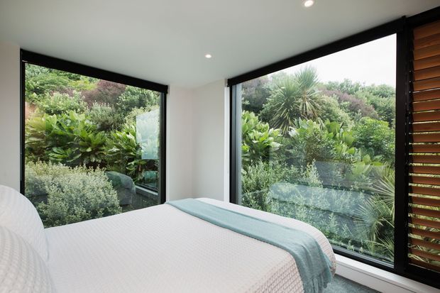 A bright, contemporary bedroom viewed from beside the bed, with floor-to-ceiling corner glazing overlooking lush garden vegetation. The room has white walls and ceiling, dark window frames, recessed ceiling lights, a pale upholstered or covered bed, and a soft blue-green throw. Timber horizontal venetian blinds cover the side window, adding warmth and privacy.