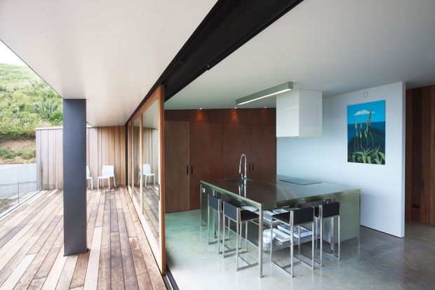 Contemporary open-plan kitchen opening onto a timber deck through full-height glazing. The kitchen features dark timber cabinetry, a large dark stone island with integrated sink and seating for several people, pale walls, polished concrete flooring, and minimalist linear lighting. A covered outdoor deck and landscaped hillside are visible beyond the glazing.