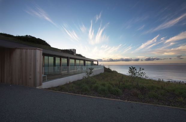 A low-profile contemporary coastal residence set directly beside a calm shoreline. The building has extensive floor-to-ceiling glazing, a long covered terrace with transparent balustrades, vertical timber-clad sections, a dark low-pitched roof, and a white rendered base. Native coastal vegetation surrounds the home, with broad water views at sunset.