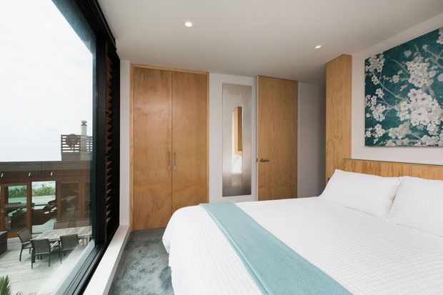 A bright, contemporary bedroom with a large bed dressed in white bedding and a pale blue runner. Full-height glazing on the left overlooks an outdoor deck and water beyond. The room includes warm timber built-in wardrobes, a timber door and headboard, a narrow full-length mirror, recessed ceiling lights, grey carpet, and a blue floral artwork above the bed.