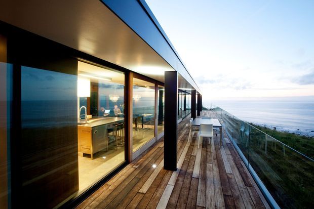 Modern coastal residence viewed from a long timber deck running alongside full-height glazed walls. The open-plan interior visibly contains a kitchen and dining area, while the deck has outdoor dining furniture and glass balustrading. The building features a crisp flat roof, dark vertical facade elements, extensive glazing, and an elevated ocean-facing setting.