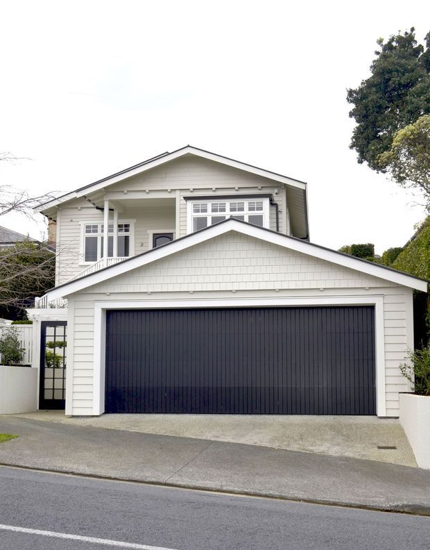 Street-facing exterior view of a two-storey light-coloured weatherboard house with a prominent attached double garage. The home has a gabled roof, white horizontal cladding, decorative shingle detailing in the garage gable, dark garage door, covered upper-level balcony or porch, and traditional residential proportions.