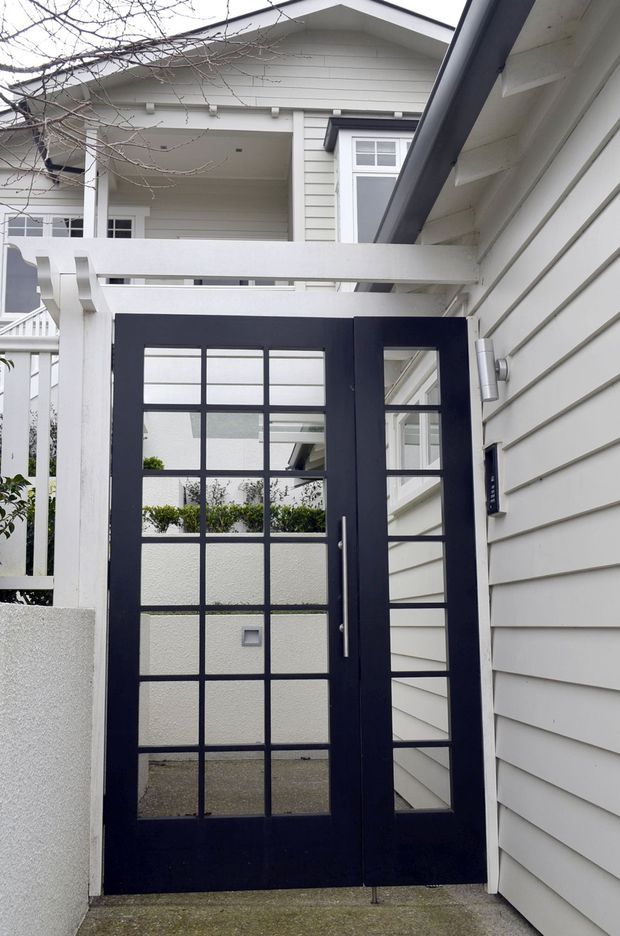A close-up exterior view of a residential entry courtyard featuring a dark navy glazed entry door with multiple rectangular panes and a matching narrow sidelight. White horizontal weatherboard cladding, white pergola beams, porch framing, and a gabled upper facade create a crisp, light-toned entrance. The image also shows a concrete-paved approach and glimpses of planted courtyard areas through the glazing.