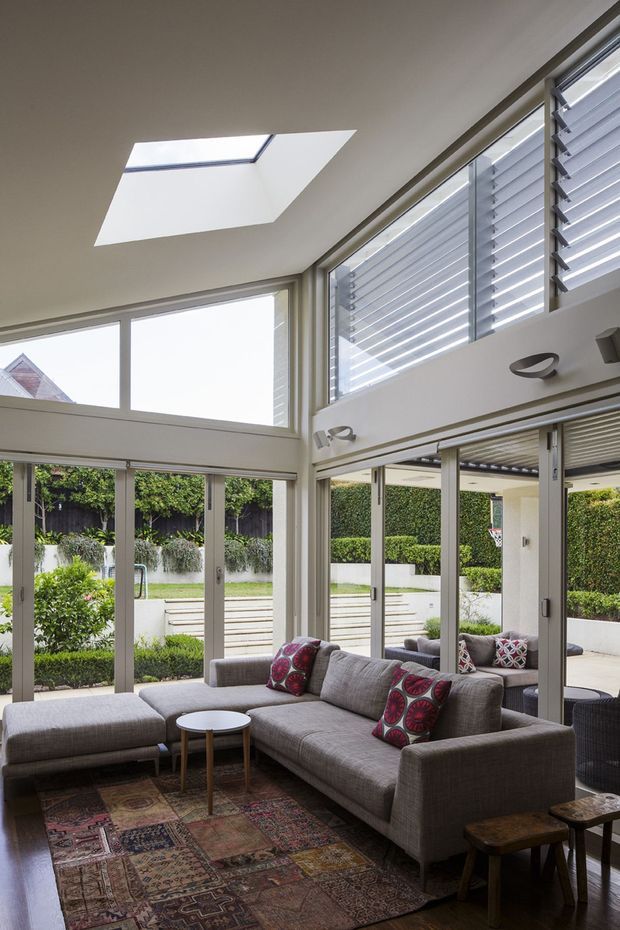 Bright contemporary living room with a large grey sectional sofa, patterned cushions, and a layered patterned rug. Floor-to-ceiling sliding glass doors open to a landscaped courtyard, while clerestory glazing, external louvres, and a large skylight provide abundant natural light. The room has a high ceiling and a strong indoor-outdoor connection.