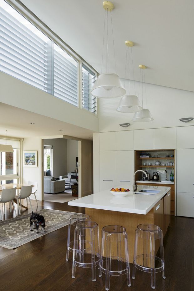 Bright, open-plan contemporary kitchen and dining area with a large white island, integrated white cabinetry, timber flooring, and transparent acrylic bar stools. A row of white pendant lights hangs beneath a high ceiling, with clerestory-style louvred windows providing abundant natural light. The dining area and adjoining living room are visible beyond the kitchen.