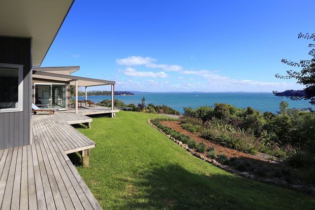 Exterior view of a contemporary coastal home with dark vertical cladding, extensive timber decking, a covered outdoor terrace, landscaped lawn and garden, and broad ocean views beyond.