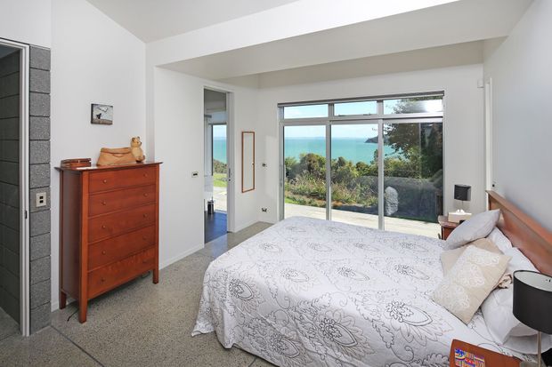 Bright contemporary bedroom with a large bed, patterned white bedding, timber headboard, tall windows and a glazed door opening to a leafy outdoor view. The room also includes carpeted flooring, a reddish timber chest of drawers, bedside tables and a large opening toward an adjoining area.