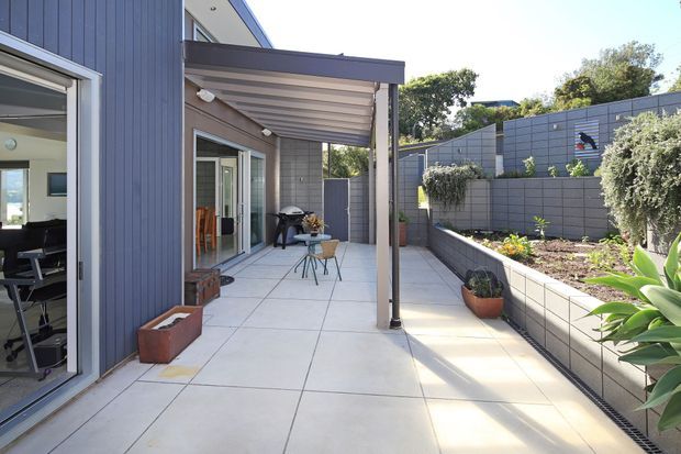 A bright contemporary outdoor patio or terrace beside a modern home, with large pale paving slabs, a covered pergola, sliding glass doors, and a raised garden bed enclosed by grey block walls and fencing. Outdoor furniture and potted plants create a functional entertaining space.