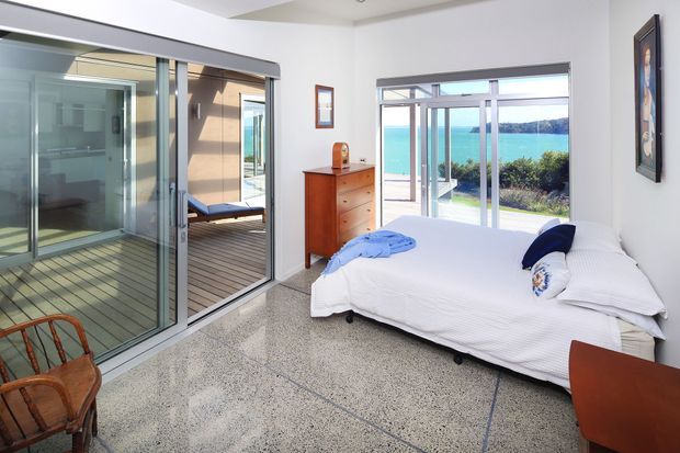 Bright contemporary bedroom with a white bed, timber chest of drawers, polished terrazzo-style flooring, and extensive sliding glass doors opening to a timber deck. The glazing provides views toward turquoise water and a distant wooded shoreline, while a small seating area is visible outside.