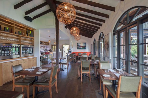A refined restaurant or café dining interior with rows of timber tables and upholstered chairs, a long built-in bar and glassware display, dark timber flooring, white walls, exposed dark timber beams, arched floor-to-ceiling windows and doors, and woven spherical pendant lights. The space has a warm, sophisticated atmosphere with views toward an outdoor terrace.