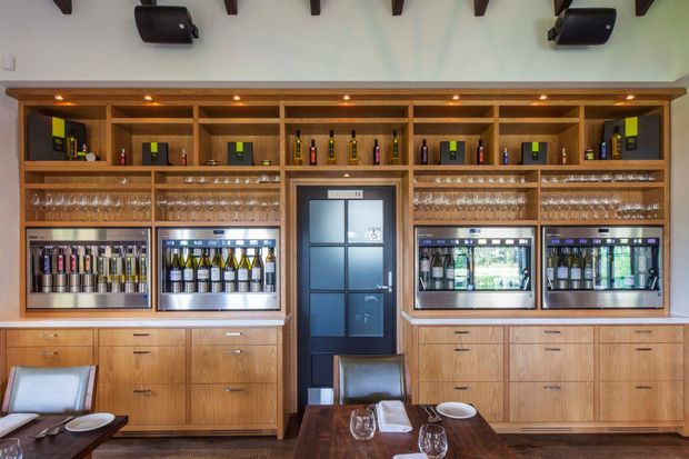 A refined commercial hospitality interior featuring a full-height timber beverage display and service wall. The built-in joinery includes illuminated bottle shelves, rows of wine glasses, under-counter wine refrigerators, and extensive timber drawers and cabinetry. A dark framed door is centered in the composition, with a dining table and chairs in the foreground and exposed dark ceiling beams above.