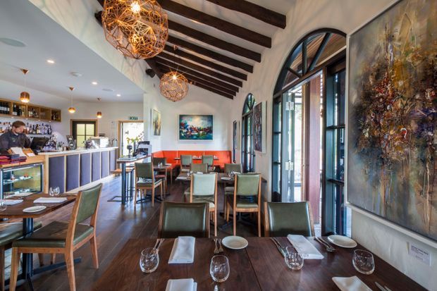 A stylish restaurant or bar interior with a long dining area, dark timber flooring, exposed dark timber ceiling beams, tall arched glazed doors and windows, a bar counter to the left, and multiple neatly set dining tables. Woven spherical pendant lights, colorful artwork, and a red upholstered banquette add visual character.
