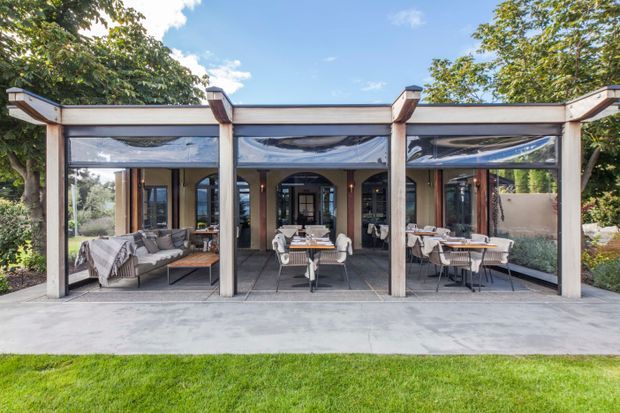 A glazed outdoor entertaining pavilion or enclosed terrace viewed from the lawn, with a flat roof supported by substantial timber posts and beams. Retractable clear screens enclose a furnished dining and lounge area, with multiple tables, chairs, and a sofa arranged on a concrete floor. Mature garden planting surrounds the structure.