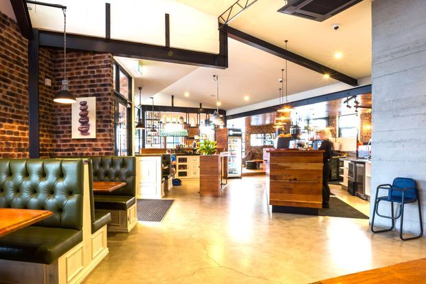 Interior of a stylish café or restaurant with exposed red brick walls, polished concrete flooring, black steel structural beams, pendant lighting, tufted green leather banquette seating, timber cabinetry, and a central wooden service counter. The space has an industrial yet warm and inviting character, with visible café equipment and dining areas.