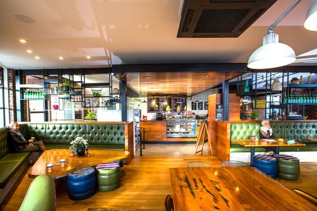 A warmly lit modern café or casual restaurant interior with extensive timber flooring, a timber-clad service counter and ceiling, black metal-framed shelving and glazing, tufted green banquette seating, colorful ottomans, and large pendant lights. The space includes dining tables, displayed bottled products, and a visible menu/service area.