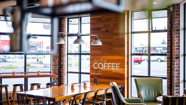A bright, industrial-style coffee shop interior with large black-framed windows, exposed brick columns, timber wall panelling, pendant lights, a long communal wooden table, bar stools, dining chairs, and a green leather armchair. The timber feature wall carries coffee-themed lettering.