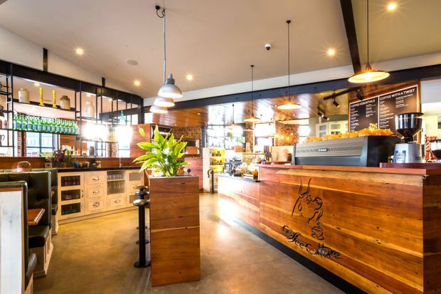 Warm, industrial-style café interior featuring a substantial timber service counter with built-in espresso equipment, hanging pendant lights, exposed brick accents, open shelving with bottles, upholstered booth seating, and polished concrete flooring.