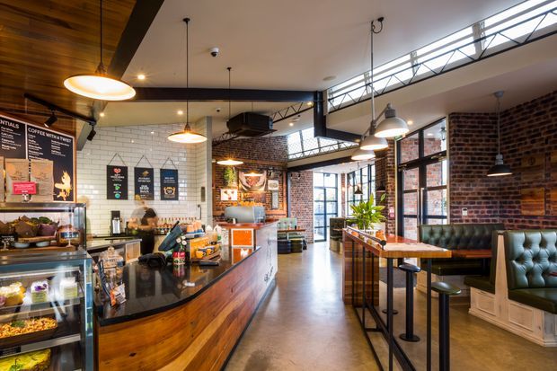 An industrial-style café interior with a long timber service counter, black stone worktops, white subway-tile walls, exposed red brick, polished concrete flooring, steel-framed windows, and suspended pendant lighting. The space includes a customer seating area with upholstered booth seating and high timber tables, plus visible café equipment and menu boards.