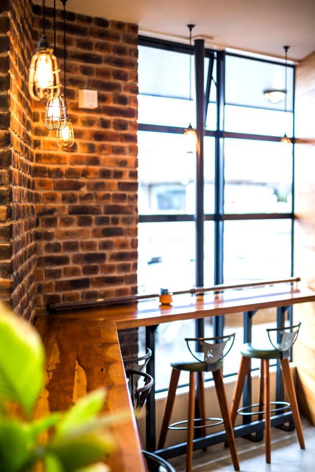 A warmly lit industrial-style interior with an exposed red-brown brick feature wall, tall black-framed windows, suspended cage pendant lights, and a long timber-topped bar counter with high stools. The space appears to be a café, bar, or hospitality venue, with timber and metal finishes throughout.