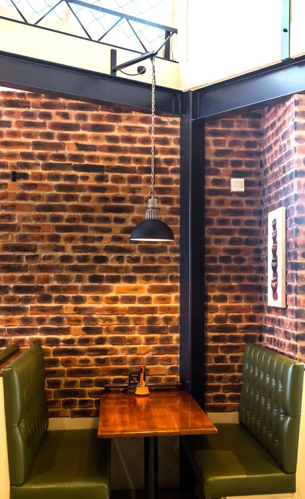 A compact industrial-style dining booth with exposed red-brown brick walls, black-painted steel structural beams, and a suspended black dome pendant light. Green upholstered banquette seating surrounds a warm timber dining table. High-level glazing or clerestory windows and visible steel bracing add an industrial character.